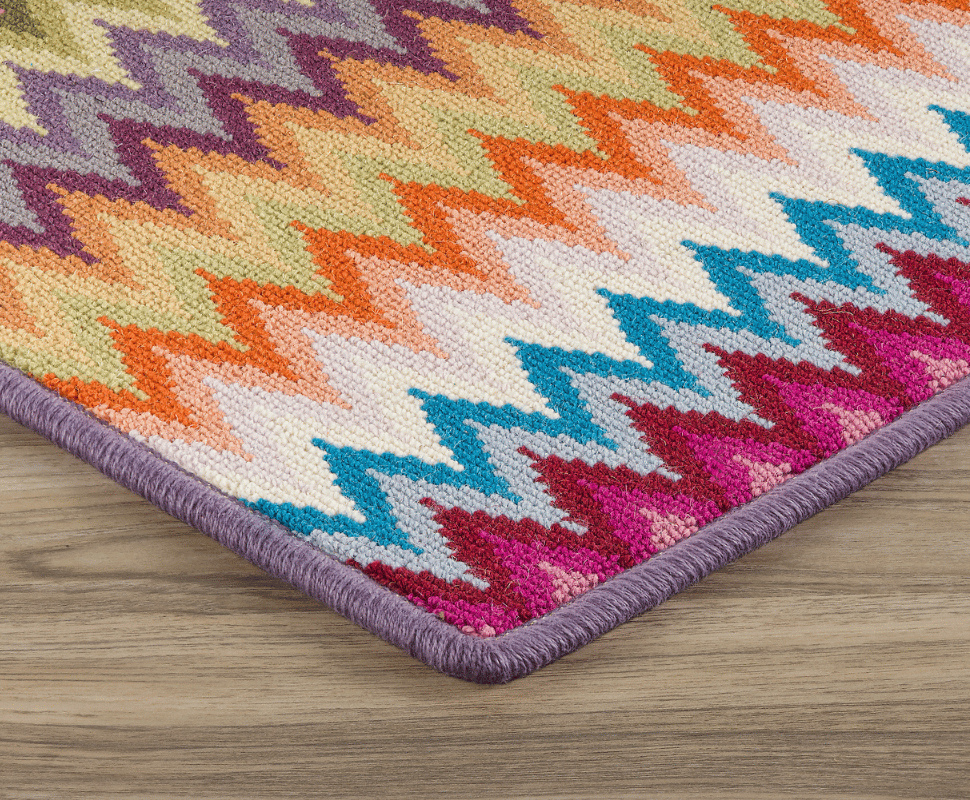 Purple bound edge bordering a vibrant multi-color flame-stitch wool loop rug on wood flooring.